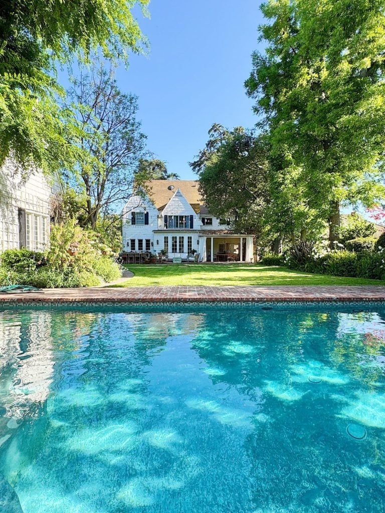 A clear swimming pool in the foreground with a large white house, green lawn, and tall trees in the background under a blue sky.