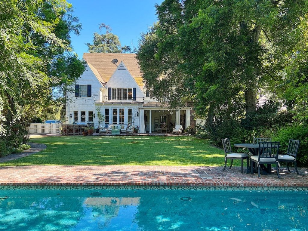 A white two-story house with a tan roof sits behind a lawn and patio, with outdoor furniture and a swimming pool in the foreground, surrounded by trees and greenery.