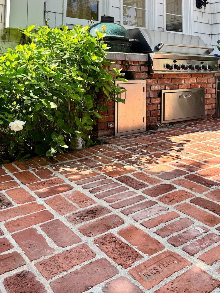 Outdoor brick patio with built-in stainless steel grill, green ceramic smoker, and blooming shrub with white flowers in sunlight.