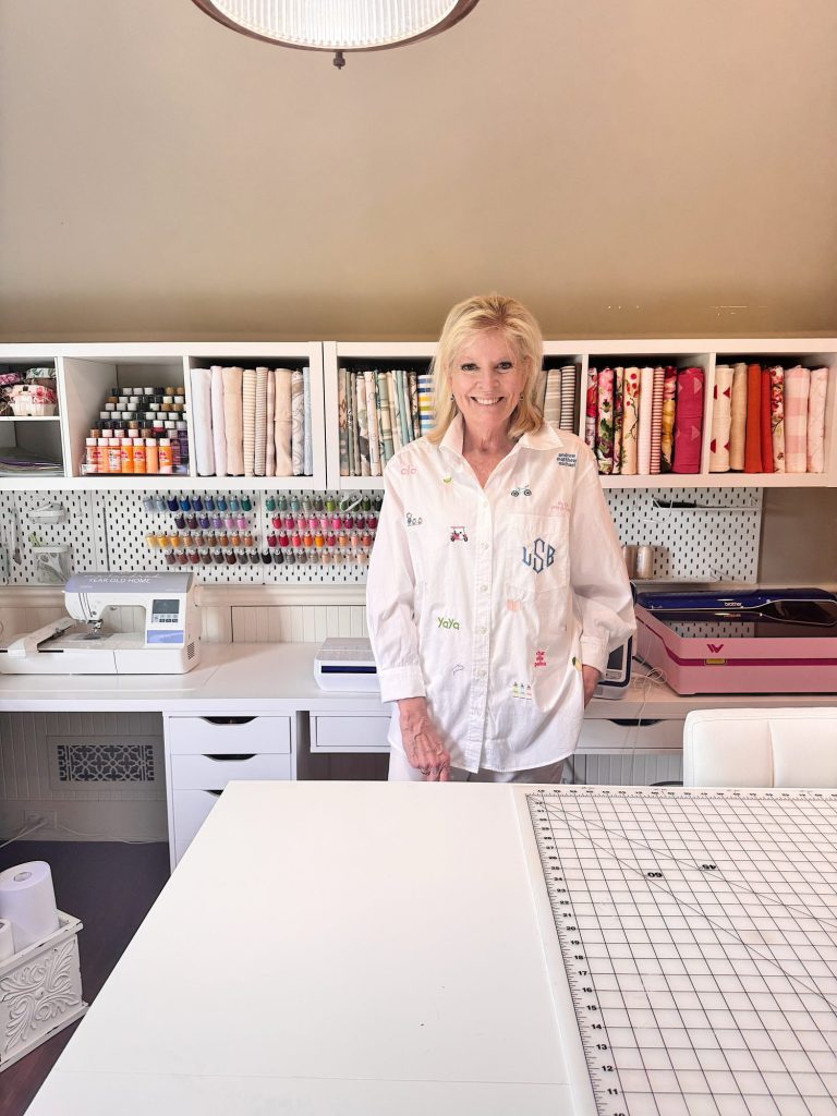 A woman stands and smiles in a sewing room with organized shelves holding fabric, thread, and art supplies. A sewing machine and cutting mat are on the white workspace.