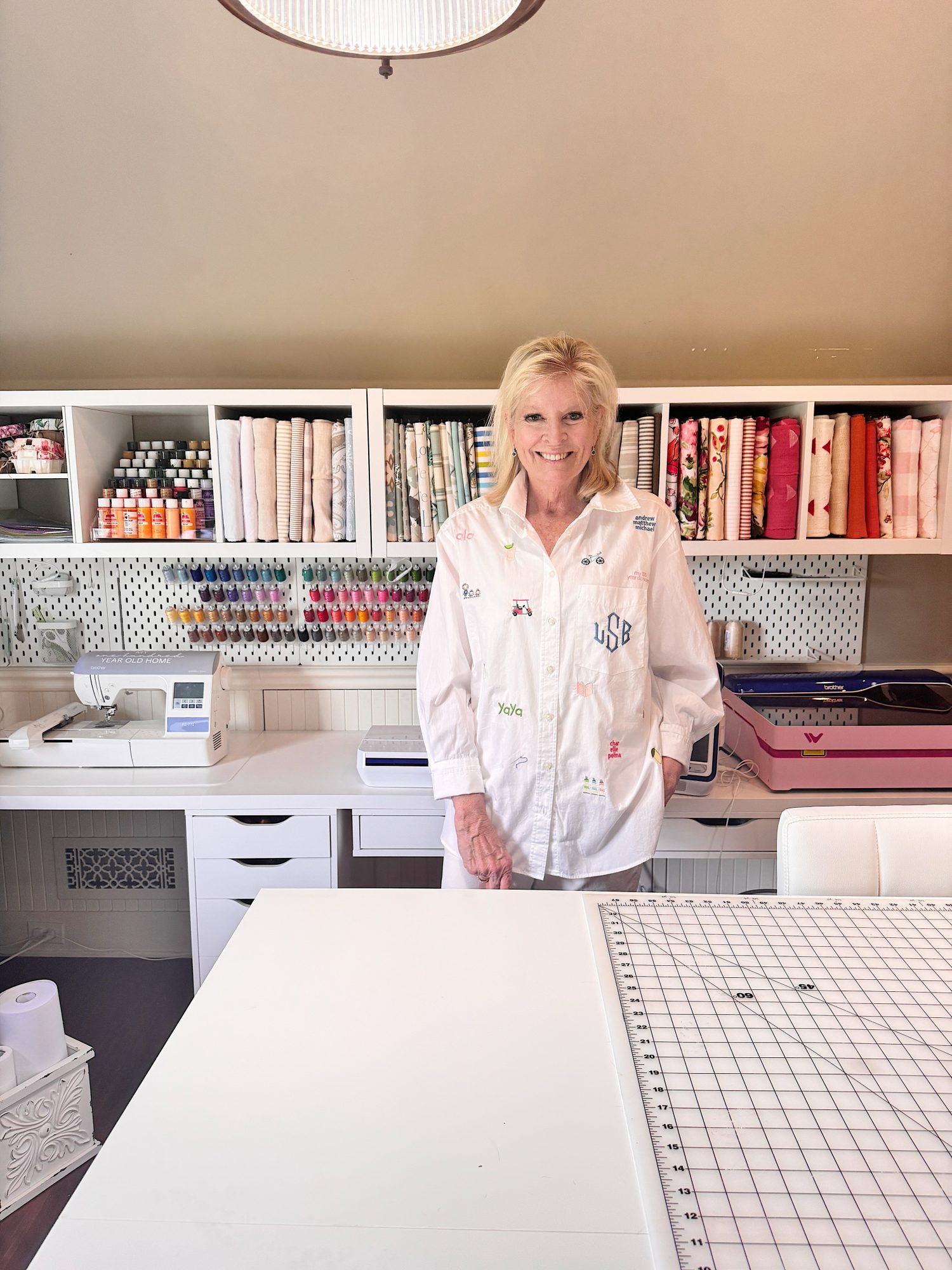 A woman stands and smiles in a sewing room with organized shelves holding fabric, thread, and art supplies. A sewing machine and cutting mat are on the white workspace.