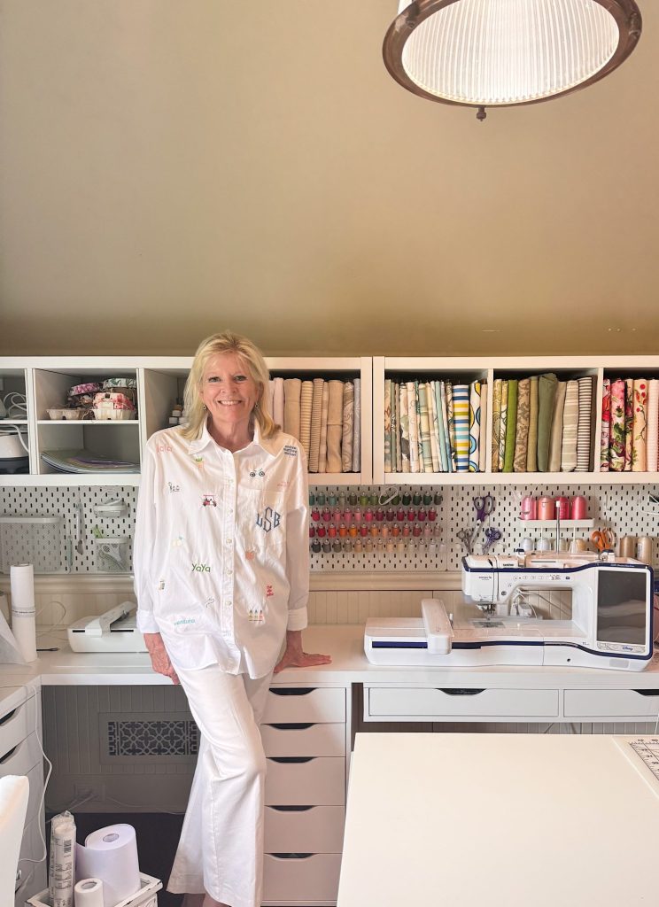 A woman in white clothing sits on a desk in a well-organized sewing room with fabric rolls, threads, and a sewing machine in the background.