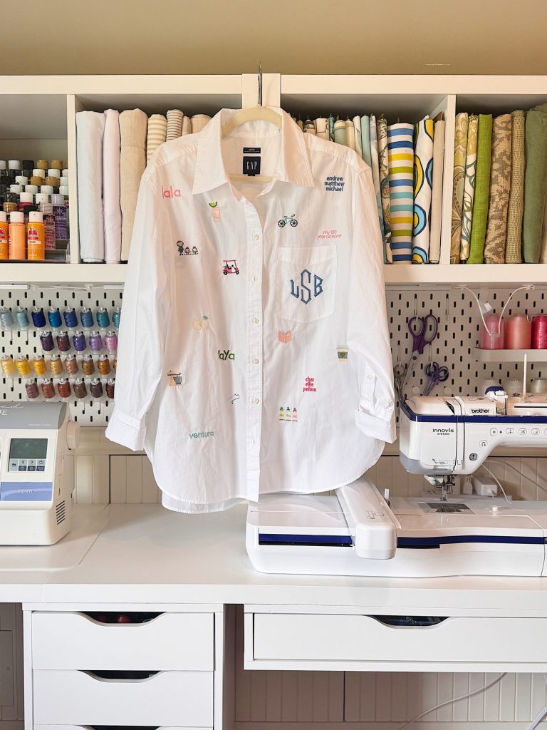 A white button-up shirt with various small embroidered designs hangs in a sewing workspace with fabric, thread spools, and sewing machines.
