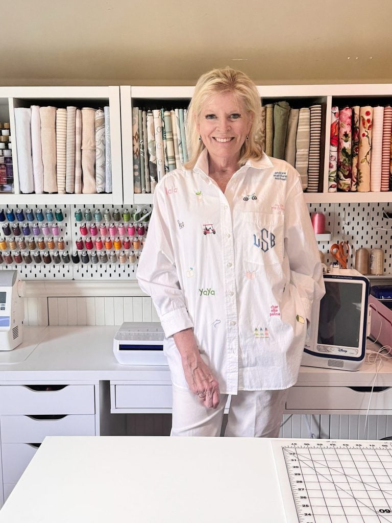 A woman in a white shirt stands in a craft room with shelves of fabric, colorful thread spools, and sewing equipment in the background.