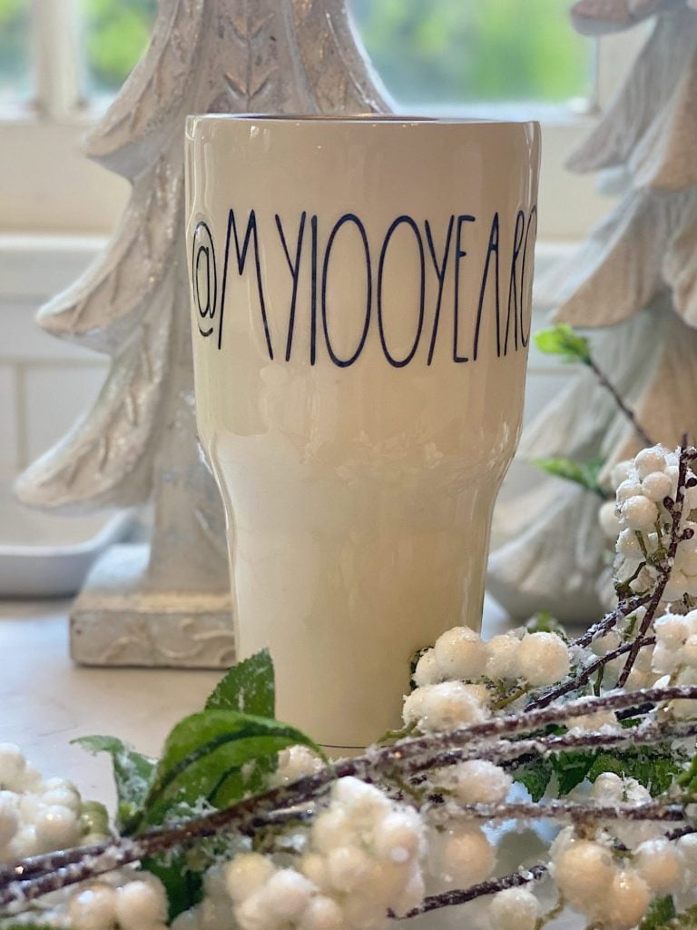 A cream-colored cup with "MY100YEARS" written in black letters, placed in front of white decorative trees and frosted faux berries.