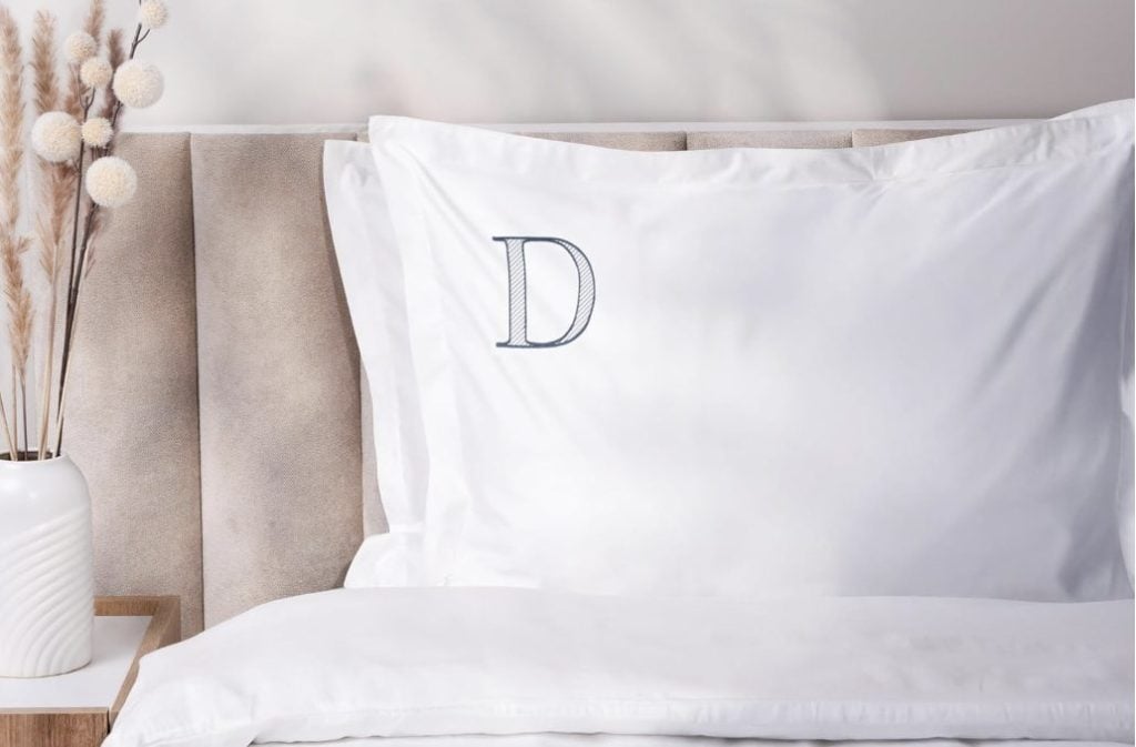 White pillow with a large embroidered letter "D" rests on a beige upholstered headboard; a vase with dried flowers sits on a bedside table.