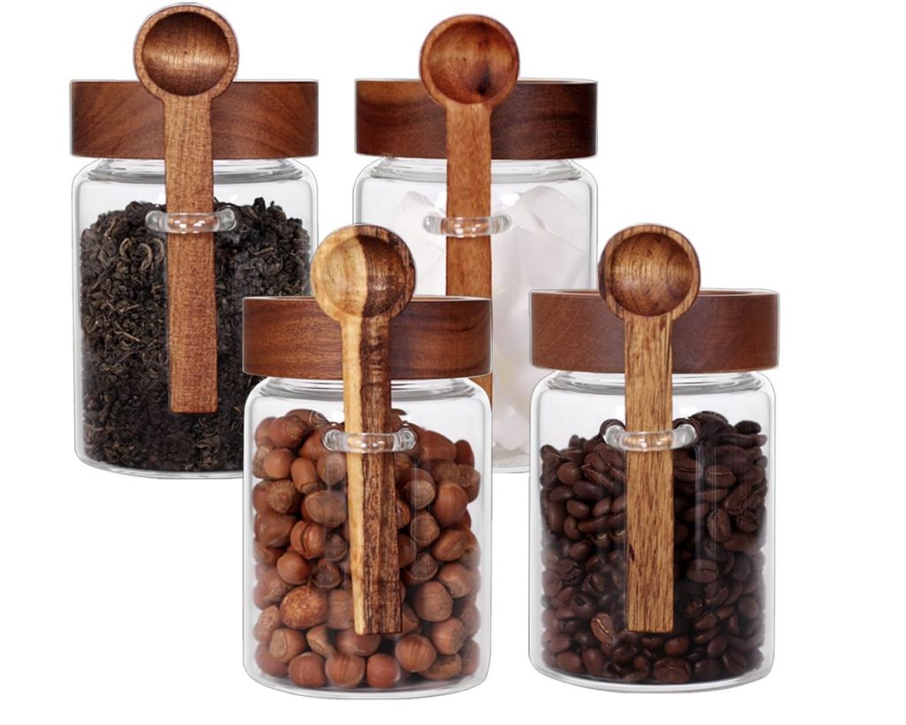 Four glass jars with wooden lids, attached wooden spoons, and filled with tea leaves, tea bags, sugar cubes, hazelnuts, and coffee beans.