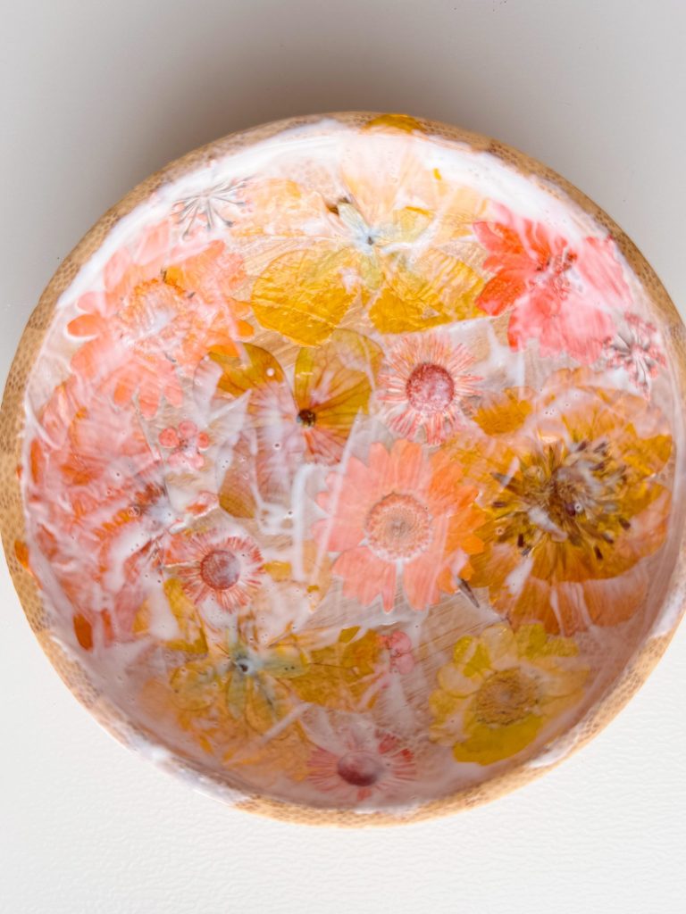 A round wooden bowl with a glossy finish, featuring embedded dried flowers in shades of yellow, orange, and pink.