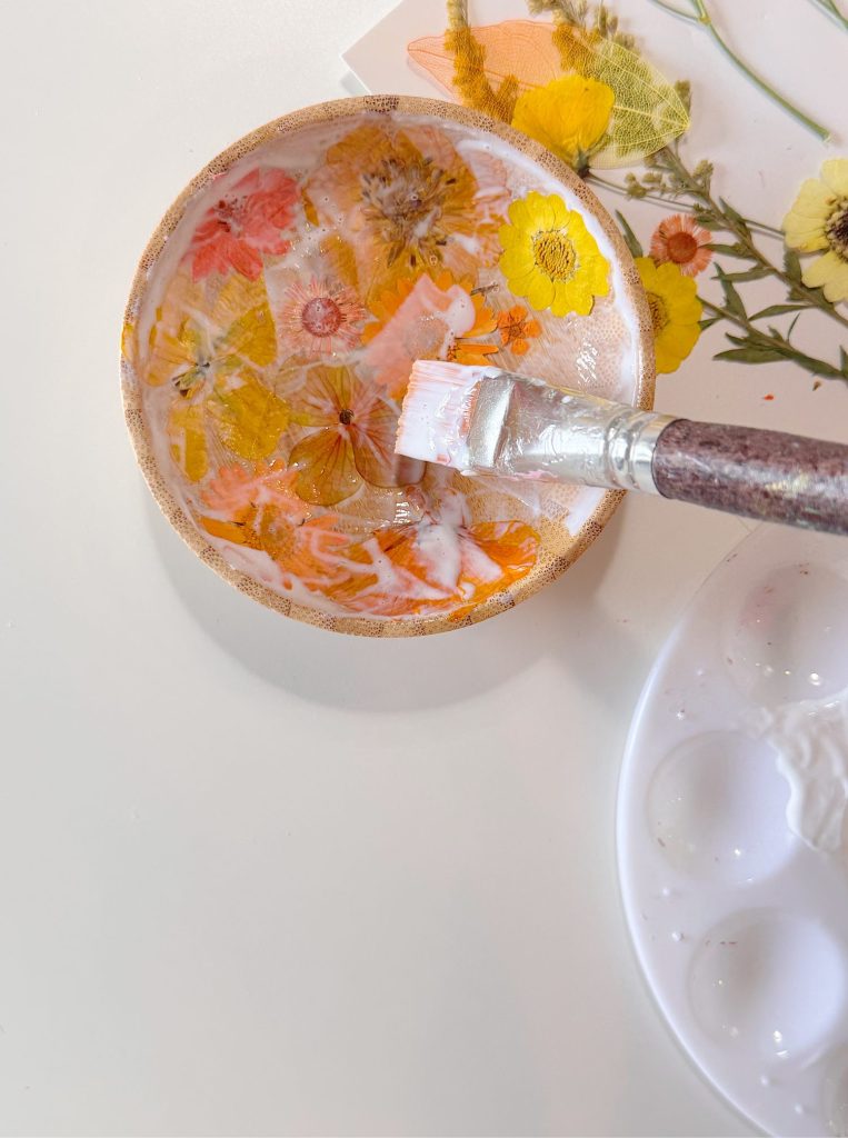 A round dish with pressed flowers being coated with a layer of clear resin using a paintbrush; palette with white paint is nearby.