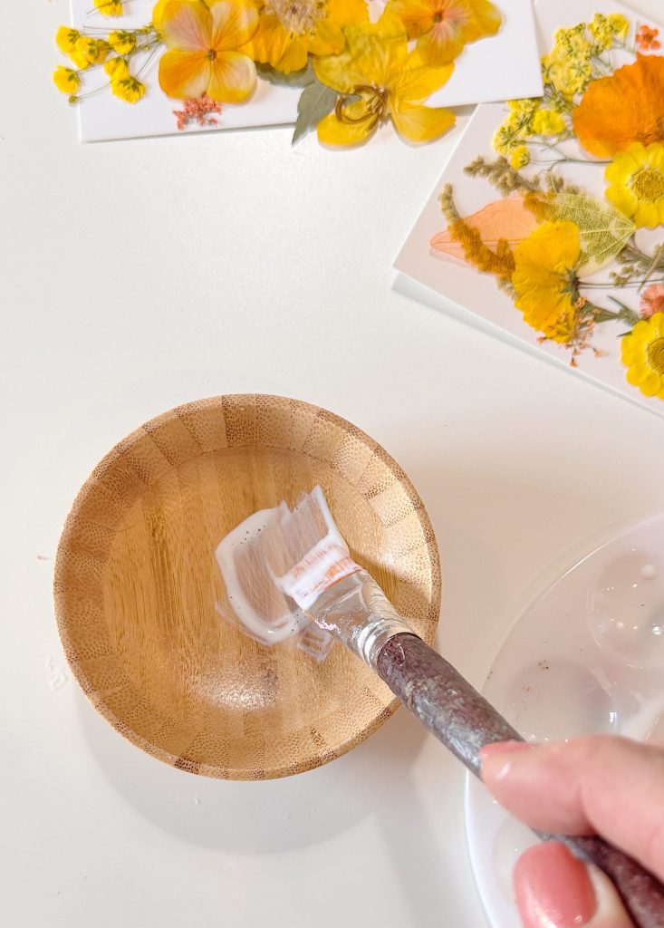 A hand holds a paintbrush with white glue over a small wooden bowl, with pressed yellow flowers and leaves arranged on cards in the background.