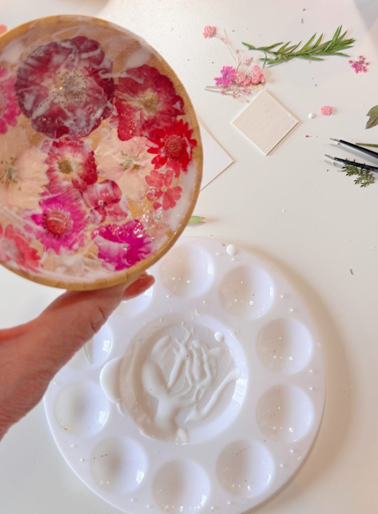A hand holds a round resin dish with embedded pink and white flowers above a white paint palette with some residue. Dried flowers and resin supplies are scattered on the table.