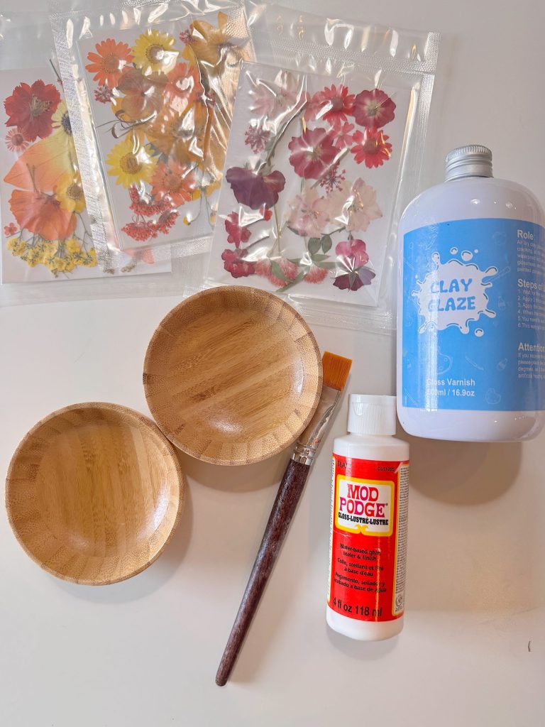 Two small wooden bowls, a paintbrush, Mod Podge, clay glaze, and several packs of pressed flower stickers are arranged on a white surface.