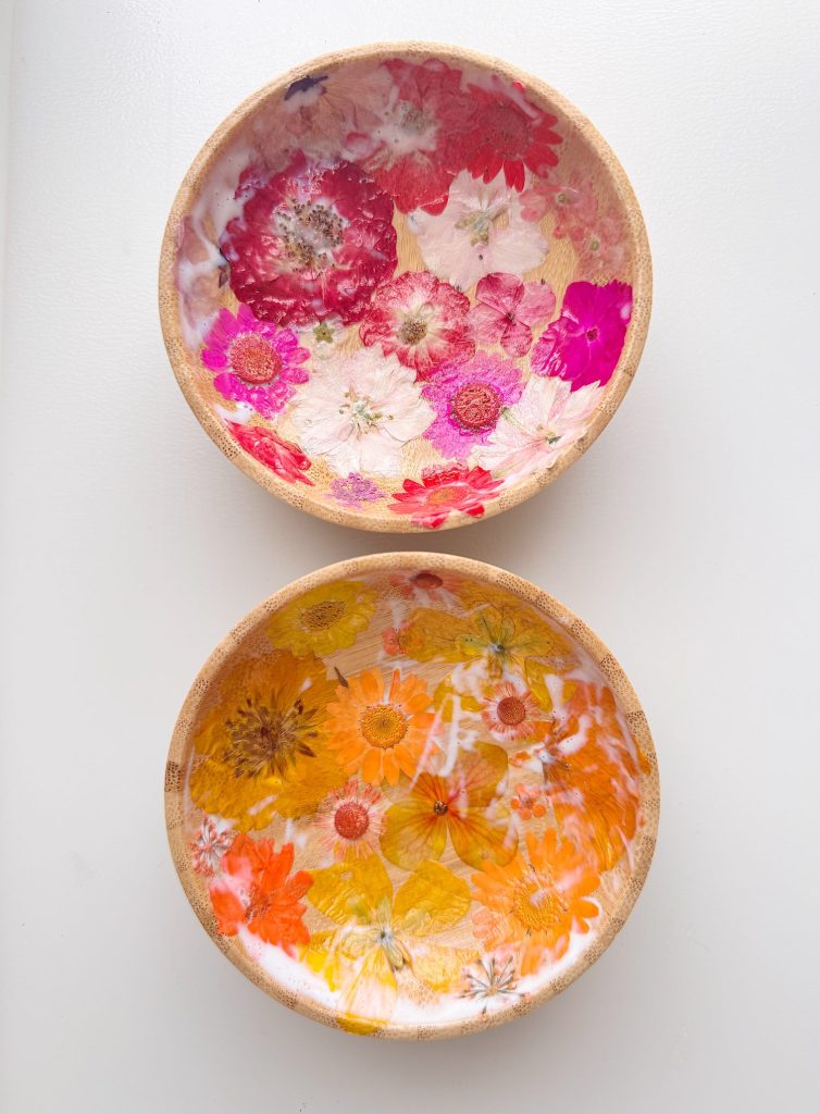 Two round wooden bowls with a glossy finish, one decorated with pink flowers and the other with yellow and orange flowers, displayed on a white surface.