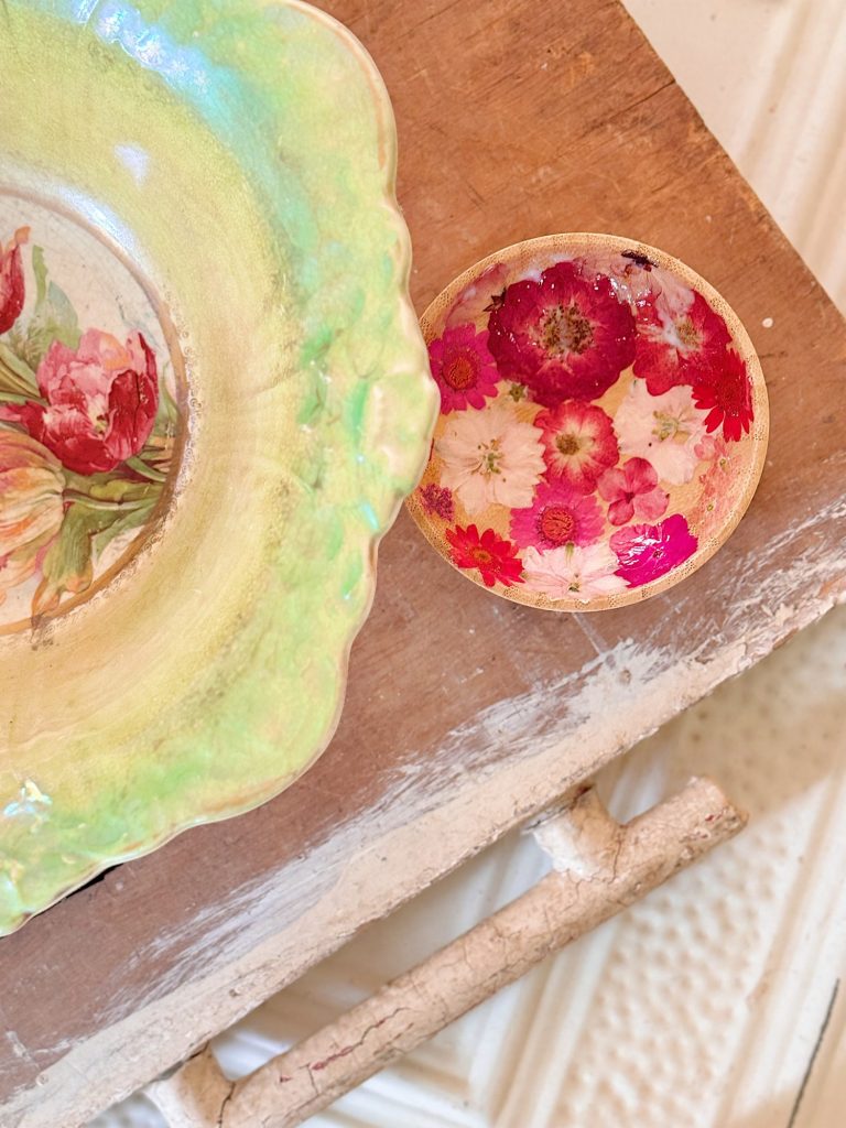 A small dish with pressed red and pink flowers sits next to a larger iridescent plate with floral designs on a wooden surface.