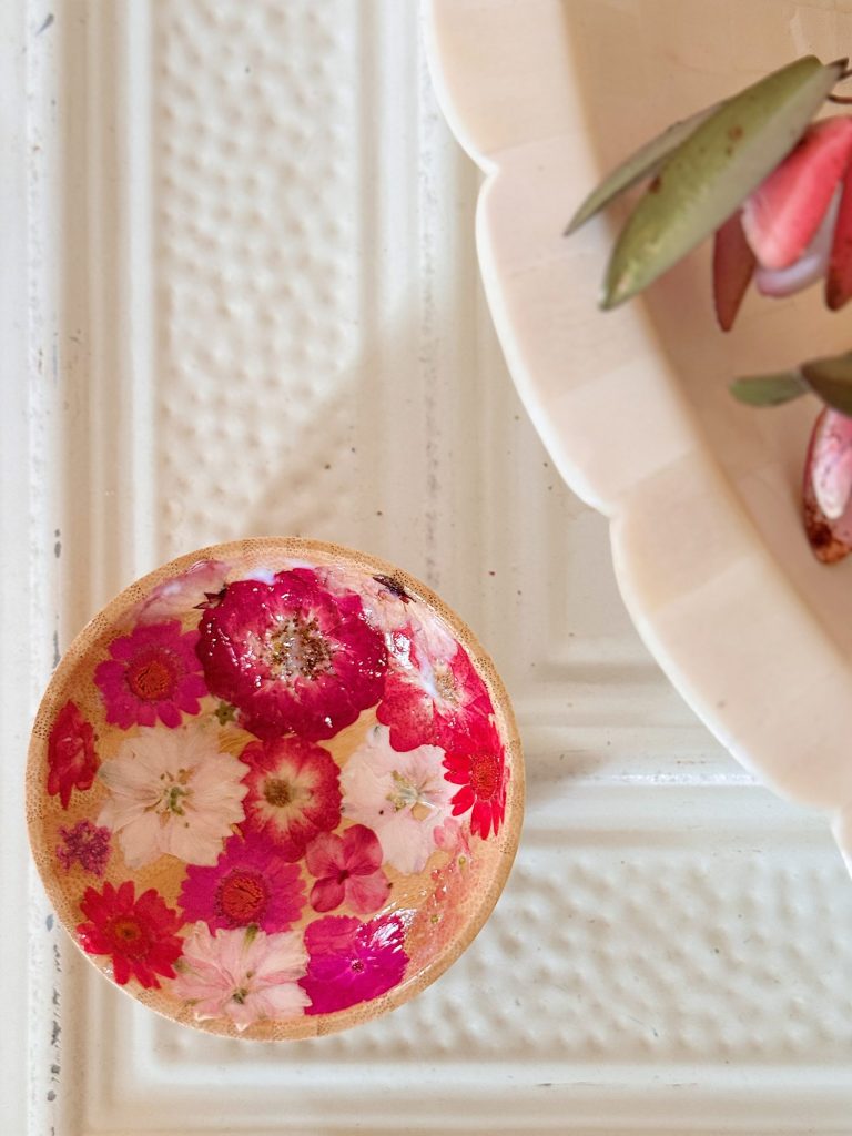 A small round dish with pressed pink and white flowers sits on a textured white surface next to part of a plate with green leaves and pink petals.