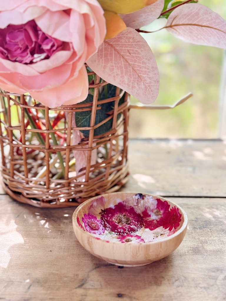 A small wooden bowl with pink flowers inside sits on a wooden table next to a wicker basket holding artificial pink flowers.