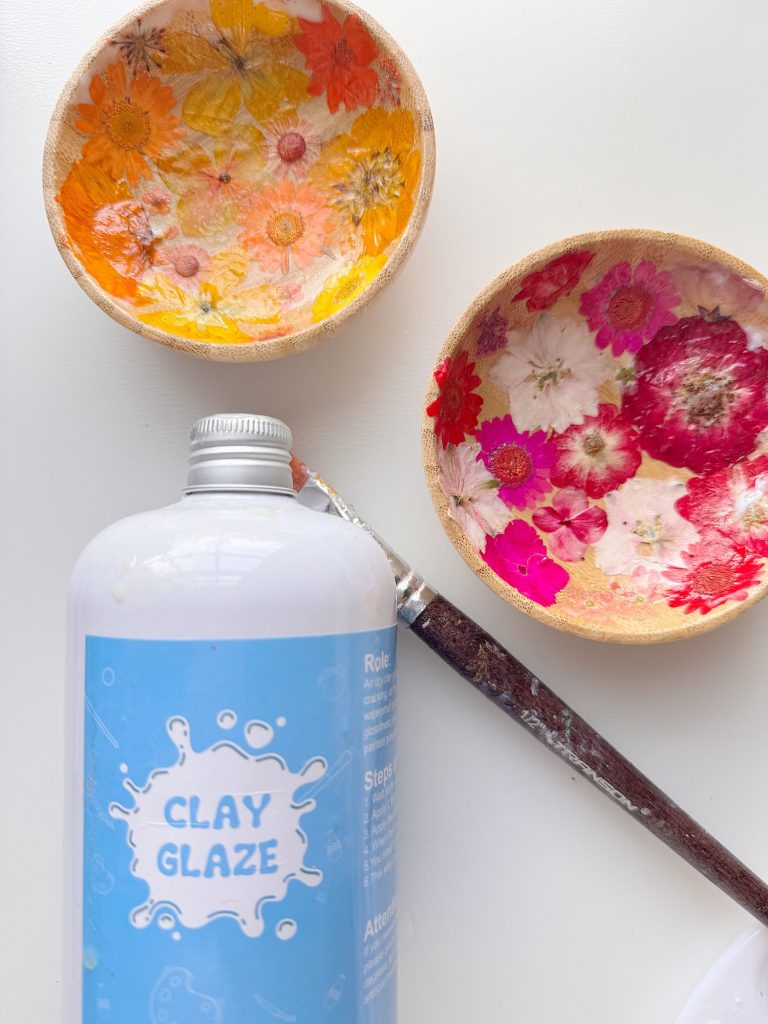 A bottle of clay glaze and a paintbrush are on a white surface next to two small decorative bowls with pressed flowers inside.