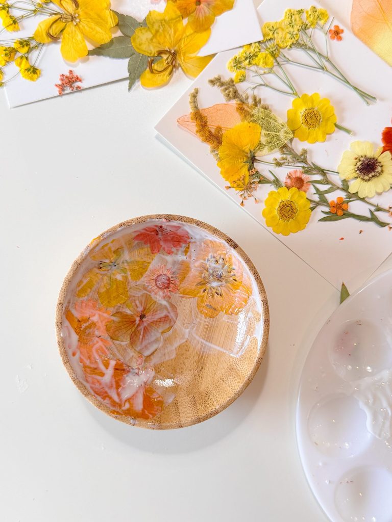 A wooden dish partially coated with resin and pressed yellow and orange flowers, surrounded by sheets with pressed flowers on a white surface.
