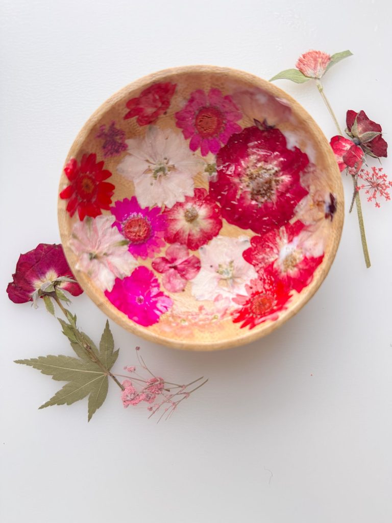 A wooden bowl decorated with pressed pink and red flowers, placed on a white surface with scattered dried flowers and leaves nearby.