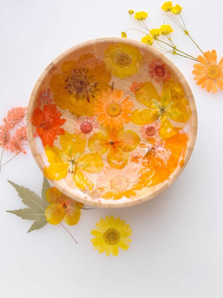 A wooden bowl decorated with pressed yellow and orange flowers, surrounded by similar flowers and a green leaf on a white surface.