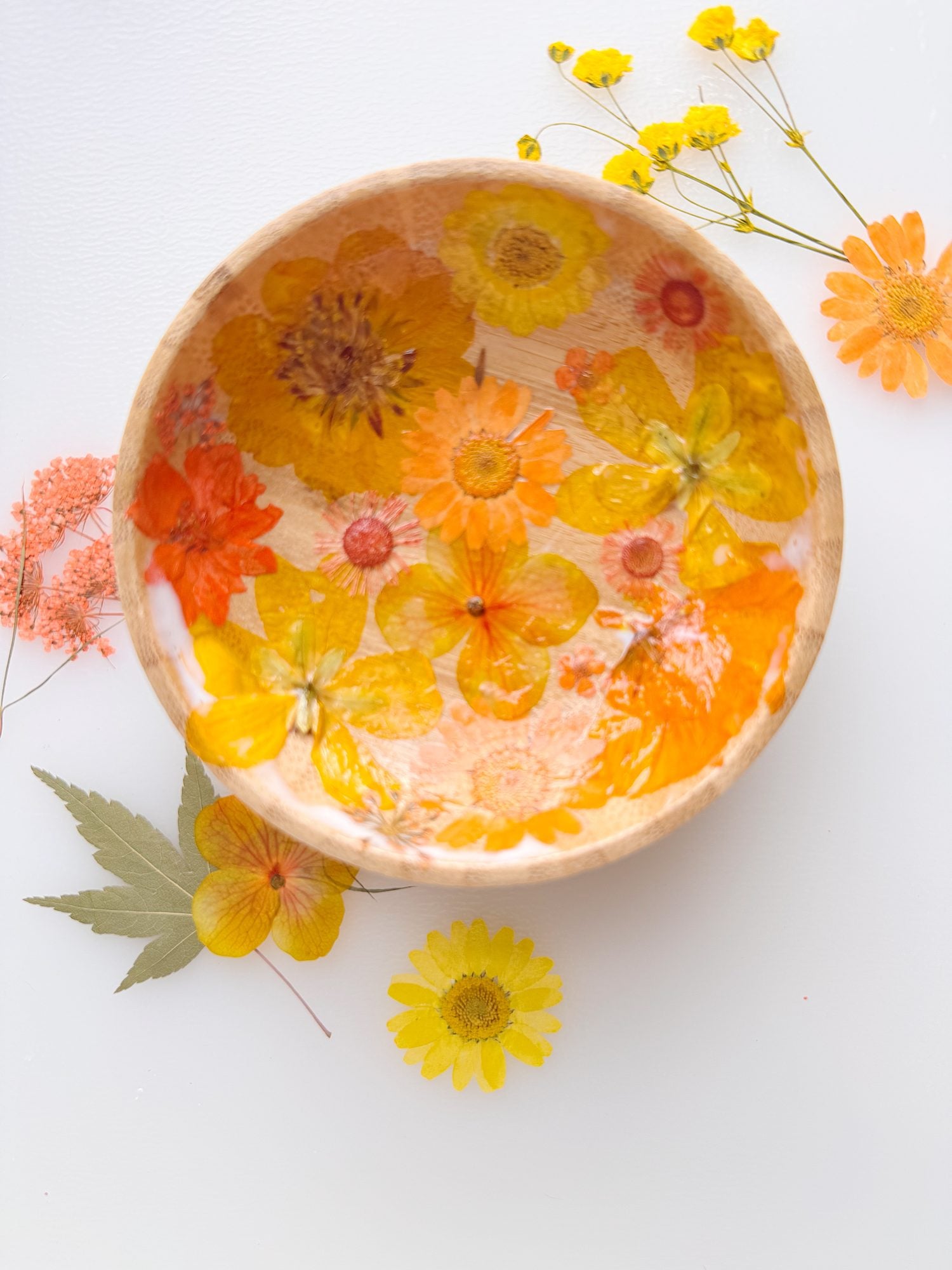 A wooden bowl decorated with pressed yellow and orange flowers, surrounded by similar flowers and a green leaf on a white surface.