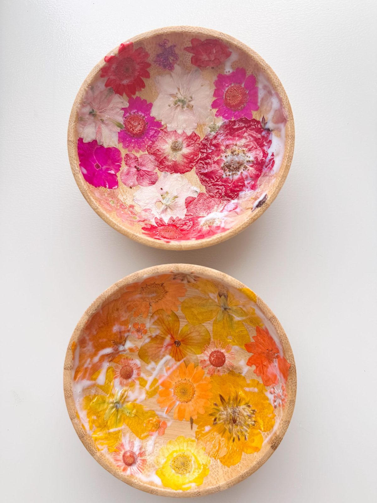 Two wooden bowls decorated with pressed flowers, one with pink and white flowers, the other with orange and yellow flowers, against a white background.