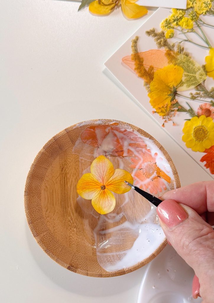 A hand uses tweezers to dip a pressed yellow flower in white glue inside a small wooden bowl, with more pressed flowers and a paintbrush nearby.