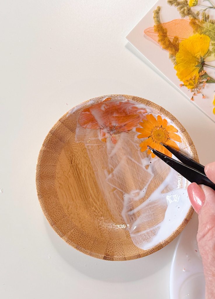 A hand uses tweezers to place a dried yellow flower covered in glue onto a small wooden bowl; more pressed flowers are visible nearby.
