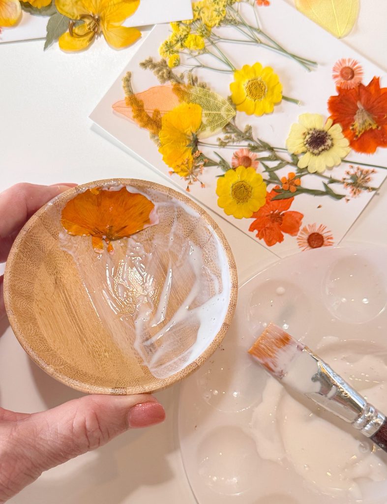 A hand holds a wooden bowl partially coated with white glue, with a pressed orange flower being applied; a brush and pressed flowers are nearby.