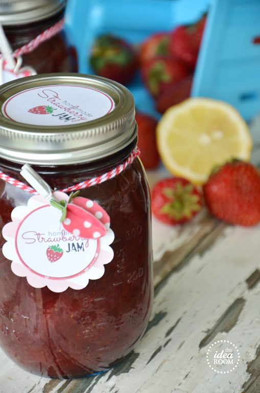 A mason jar of homemade strawberry jam with a decorative label sits on a rustic table, surrounded by fresh strawberries and a sliced lemon.