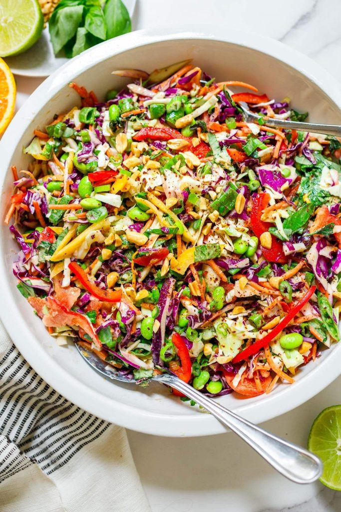 A large bowl of colorful vegetable salad with shredded cabbage, carrots, red bell pepper, edamame, herbs, and seeds, with a spoon for serving.