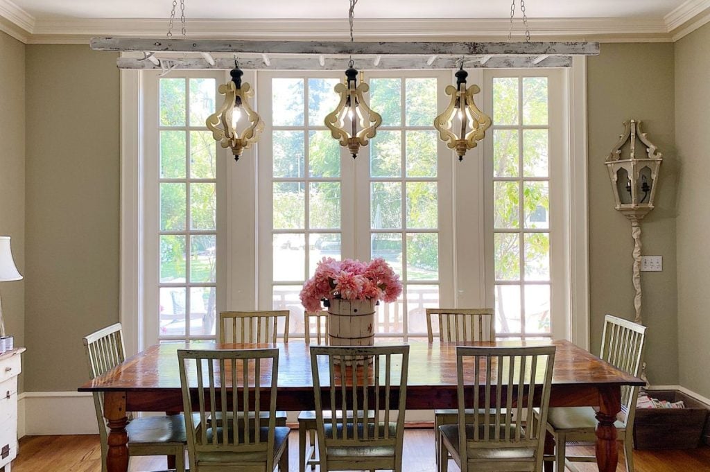A wooden dining table with six chairs sits in front of large French windows; three decorative pendant lights hang above and a vase of pink flowers is centered on the table.