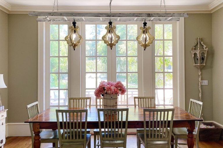 A wooden dining table with six chairs sits in front of large French windows; three decorative pendant lights hang above and a vase of pink flowers is centered on the table.