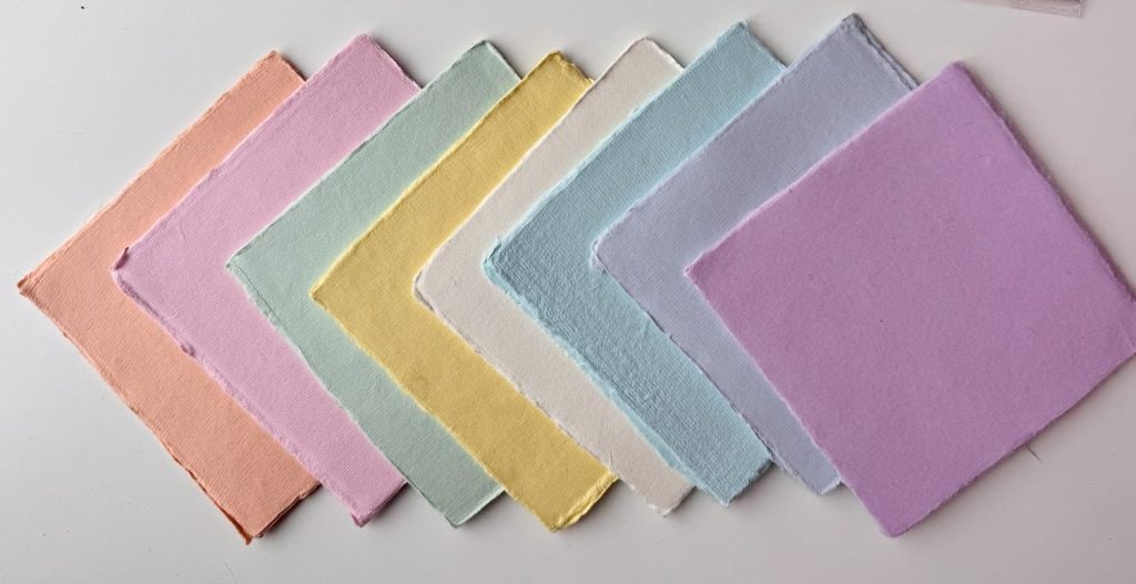 Eight square sheets of textured paper in pastel colors&mdash;peach, pink, green, yellow, cream, blue, light blue, and lavender&mdash;are arranged in a diagonal row on a white surface.