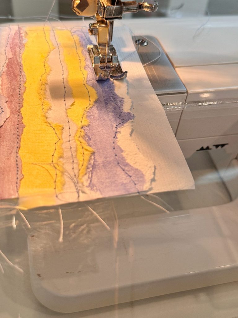 A sewing machine stitching over a piece of paper with colorful, wavy lines in purple and yellow watercolor.