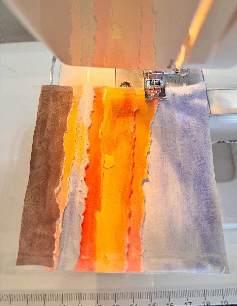 A sewing machine stitches together strips of torn, brightly colored paper in shades of brown, orange, yellow, and blue.