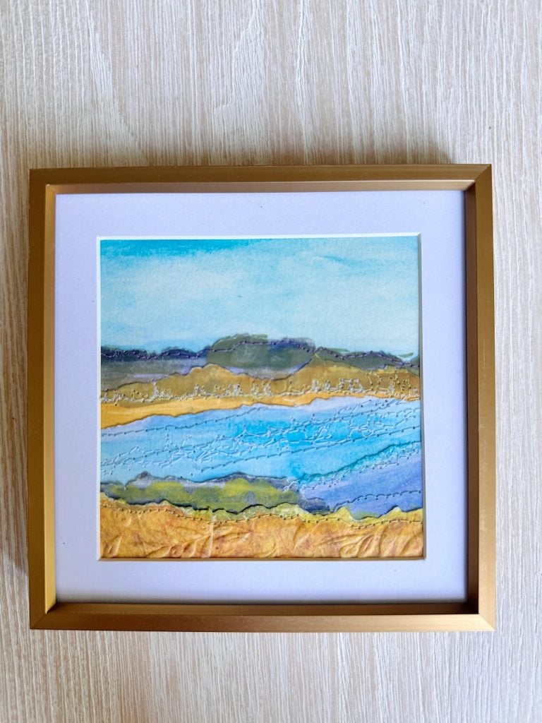 Framed abstract landscape artwork featuring layered colors and textures depicting water, land, and sky, set against a light wood background.