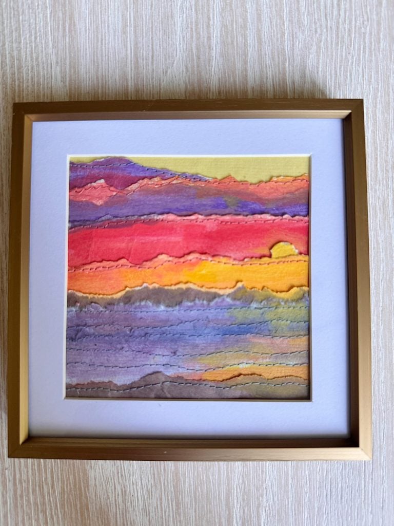 Framed artwork featuring layered, torn paper strips in various colors resembling a sunset, displayed on a light wood surface.