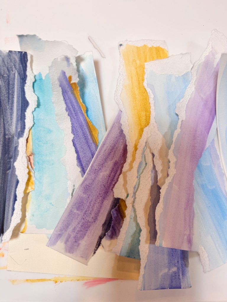 Torn strips of watercolor-painted paper in pastel colors, including blue, purple, and yellow, scattered on a white surface.