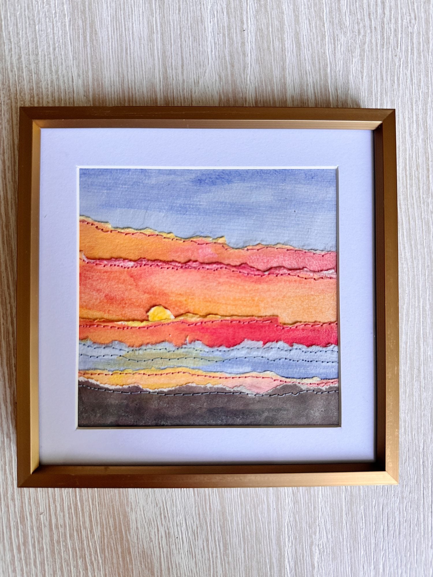 A framed artwork features layered, textured strips in sunset colors with a small sun partially visible near the horizon on a wooden surface.