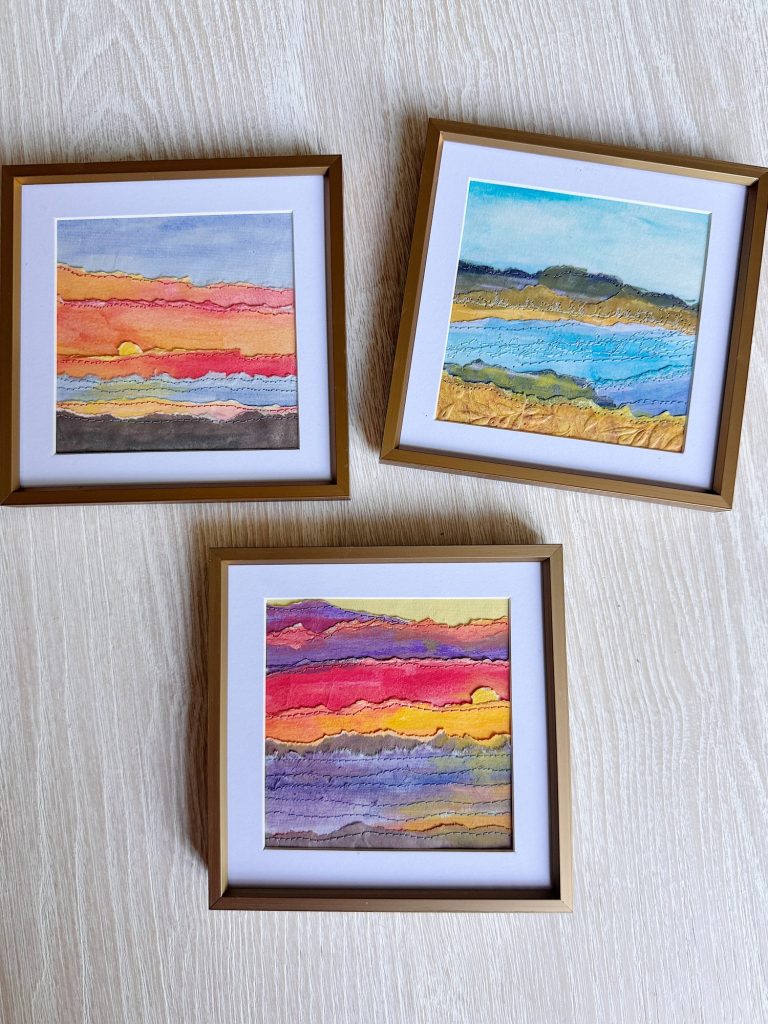 Three framed abstract landscape paintings with horizontal layers of vibrant colors are arranged on a light-colored wooden surface.