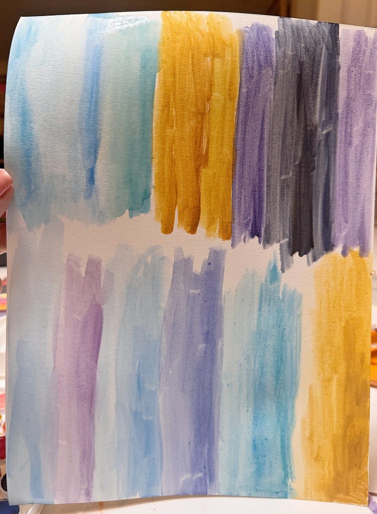 A hand holds a sheet of paper with two rows of watercolor swatches in shades of blue, purple, and yellow.