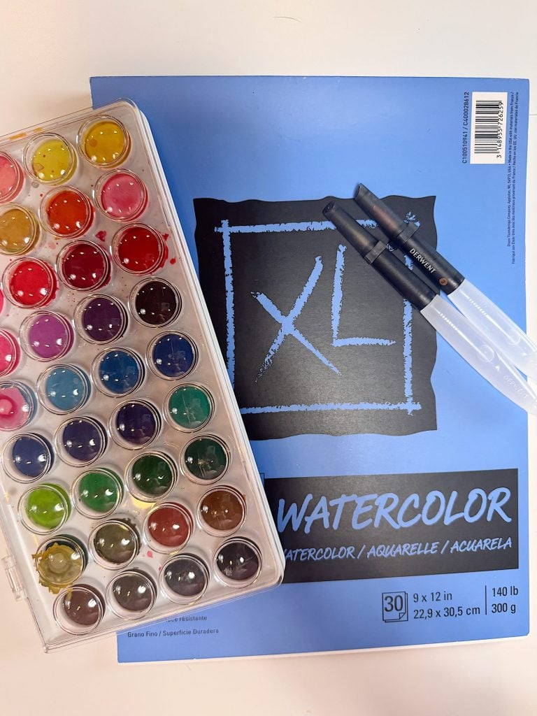 A watercolor paint set, two water brushes, and a blue XL watercolor pad are arranged on a white surface.