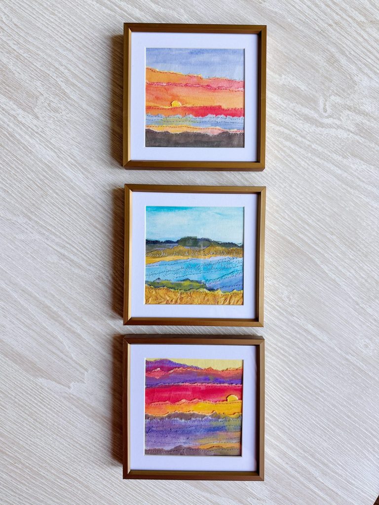 Three framed abstract landscape paintings in gold frames are displayed vertically on a light wood wall. Each painting features vibrant, layered colors.