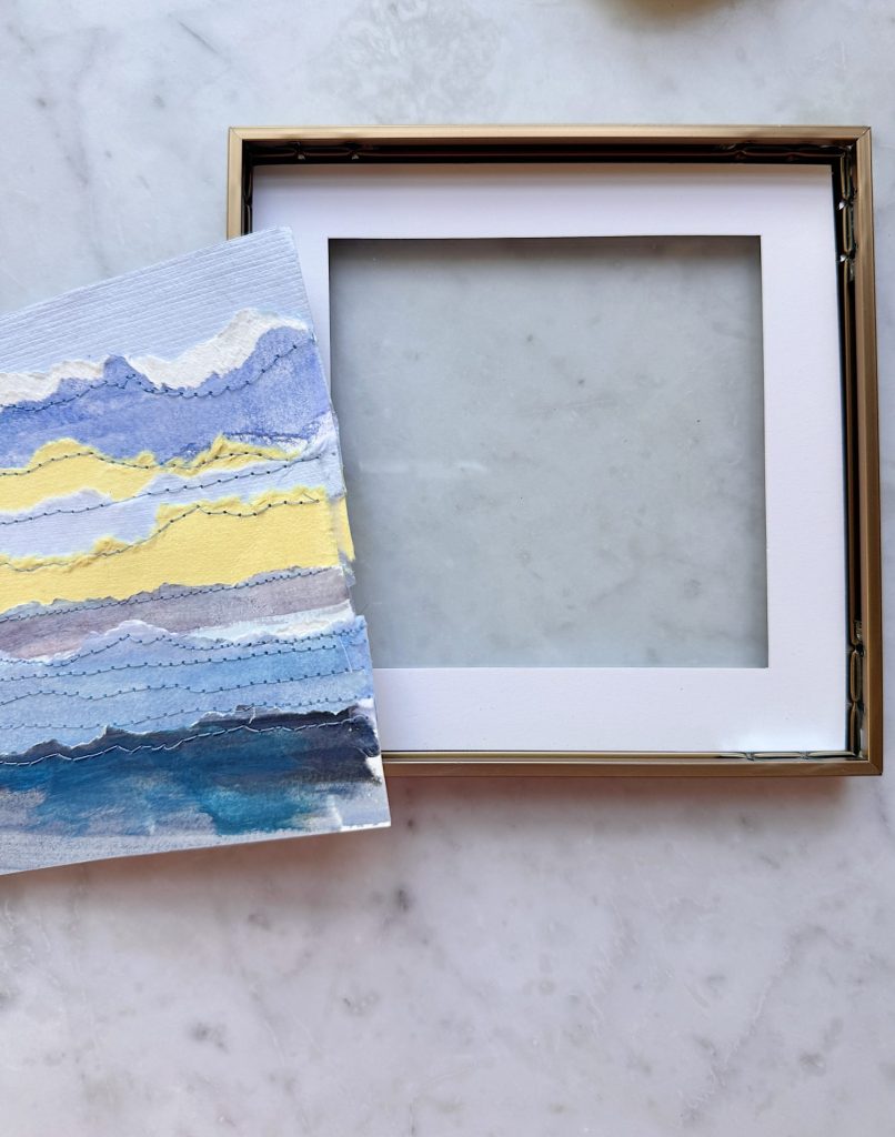 A square picture frame with a white mat lies open on a marble surface next to a torn-edge abstract painting in blue, yellow, and purple tones.