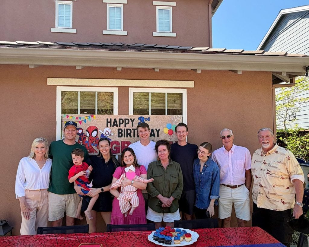 A group of eleven people, including adults and children, pose together in front of a house with a "Happy Birthday" banner and a table with cupcakes.
