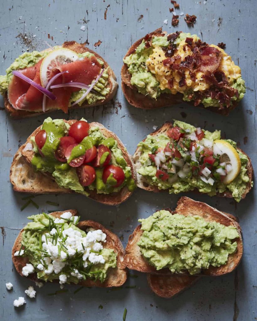 Six slices of toasted bread topped with various avocado-based ingredients, including tomato, egg, bacon, cheese, prosciutto, and fresh vegetables, arranged on a rustic blue surface.