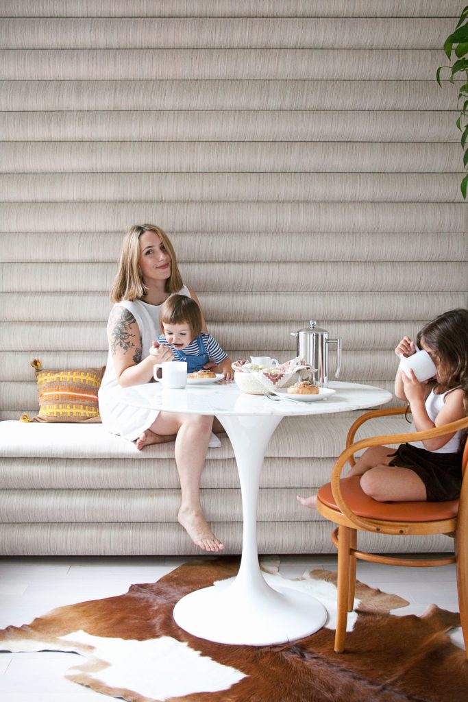 A woman and two young children have breakfast at a round marble table, with pastries, coffee, and mugs, in a bright modern room with neutral decor.