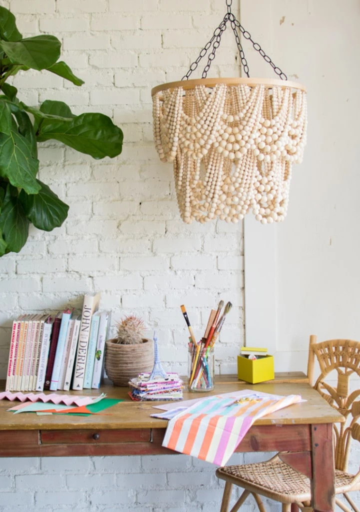 A wooden chandelier with beaded details hangs above a desk with books, art supplies, and colorful fabric, next to a woven chair and leafy plant against a white brick wall.