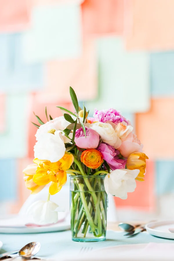A clear glass vase filled with assorted colorful flowers sits on a table set with plates and cutlery; a pastel-colored background is visible.